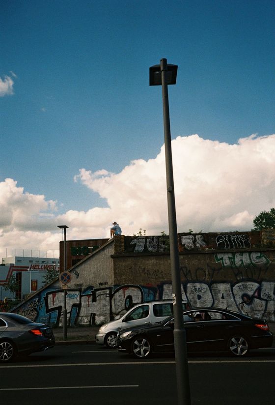 Fotografie: Parkendes Auto vor einer Mauer mit Graffiti. Auf der Mauer sitzt eine Person.