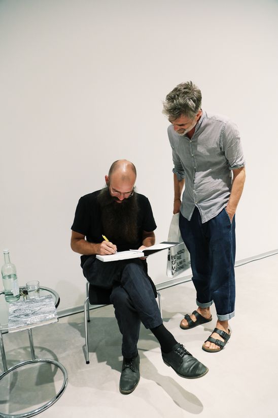 One person is sitting on a chair signing a catalogue next to another person standing.