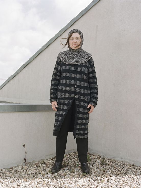 Foto: Käthe Kruse steht in kariertem langem Kleid über einer schwarzen Hose und einer Art Kragen mit Kapuze aus feinen Ketten vor einer gräulichen Wand.