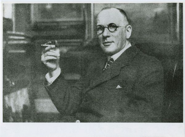 Black and white portrait of Rudolf Levy wearing a suit and holding a cigarette in his right hand. He is looking directly into the camera through round glasses.