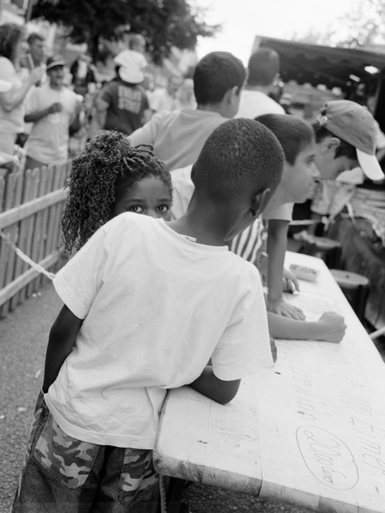 Schwarz-weiß-Fotografie: Im quadratischen Format zeigt die Fotografie mehrere Personen, die vorwiegend mit dem Rücken zur Kamera stehen, während eine junge, weiblich gelesene Person direkt in die Kamera blickt.
