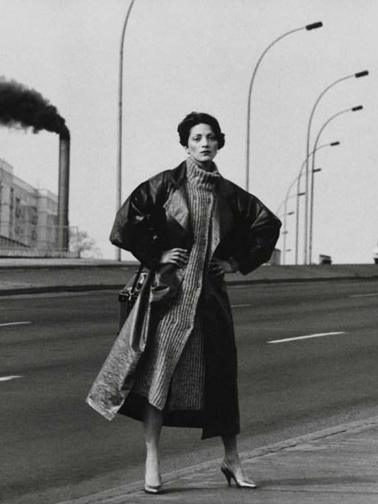 Black photograph of a woman in a coat, hands on hips, standing on a multi-lane road. In the background, streetlights and a tower from which dark smoke rises.