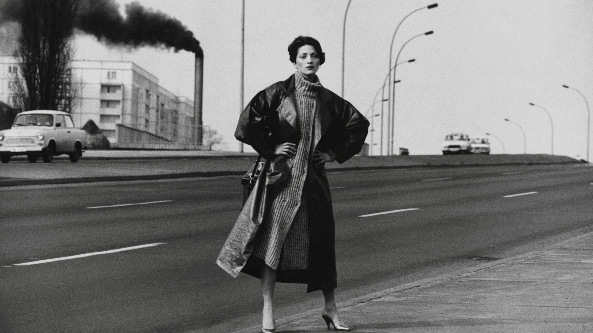 Schwarz-Fotografie einer Frau mit Mantel, die die Hände auf die Hüften gestützt, auf einer mehrspurigen Straße steht. Im Hintergrund Straßenlaternen und ein Turm aus dem dunkler Rauch aufsteigt.