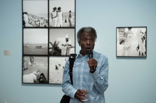 Photo: Akinbode Akinbiyi stands in the exhibition space in front of black and white photographs on a pastel blue wall and holds a microphone in his hand.