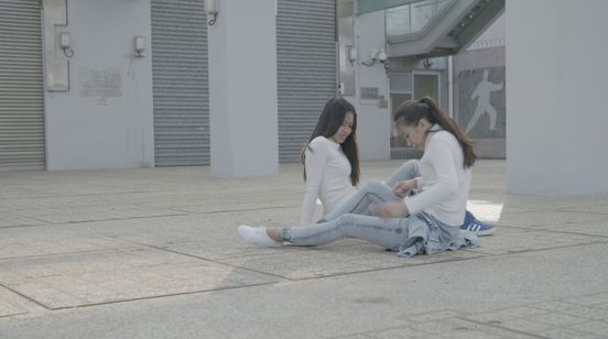 Filmstill: Zwei Personen in heller Kleidung sitzen einander zugewandt auf dem Boden zwischen zwei Gebäuden.