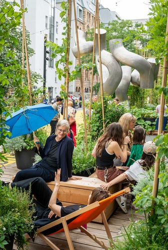 Artistic garden installation “Light and & Shadow”, atelier le balto, 2025 Photo: People sit in deckchairs and on stools on a wooden platform between large planters and a silver sculpture.
