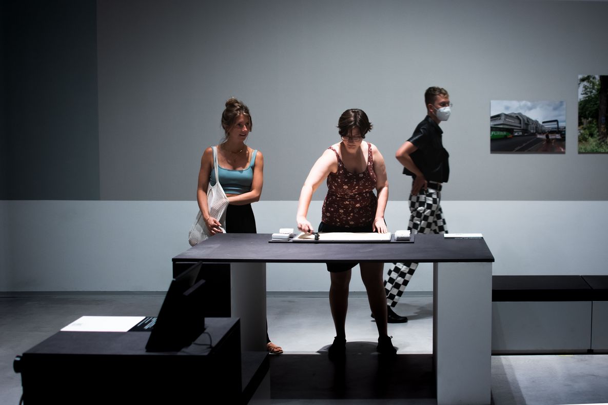 Foto: Im Ausstellungsraum steht eine Person an einem Tisch. Sie fährt mit einer Rolle über einen sandigen Untergrund.