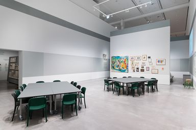 Photo: A large room with two big group tables and many chairs. On a free-standing wall hang many painted pictures and drawings.