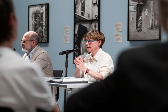 Photo: Katia Reich and Thomas Köhler sitting on a table at the press conference. Behind them are black and white photographs by Akinbode Akinbiyi.