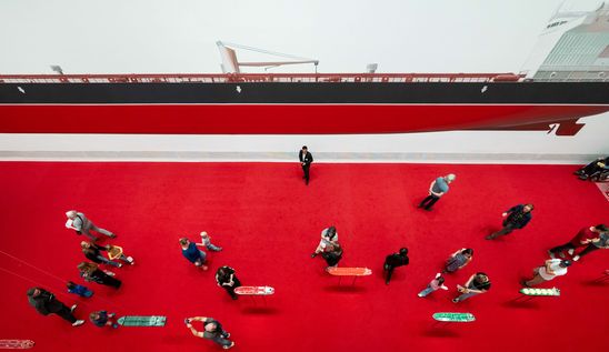 Photo: View of the exhibition room with its bright red carpet, a large reddish, shiny sculpture on the wall and a large painted oil tanker.