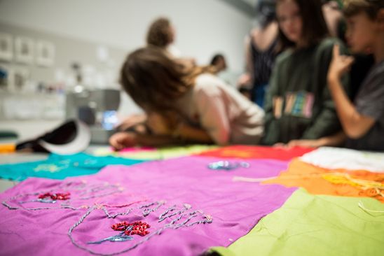 Photo: Close-up of a colourful textile work, partly embroidered with colourful beads. The background shows blurred people.