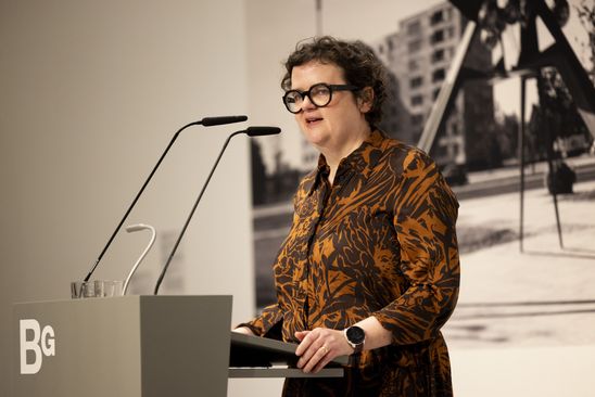 Foto: Ilka Voermann steht an einem Redner*innenpult. Im Hintergrund ist eine schwarz-weiß Foto-Tapete einer Skulptur von Hans Uhlmann am Hansaplatz in Berlin zu sehen.