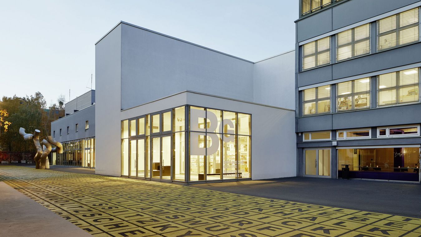 Photo: Building with a flat roof and glass front displaying the logo of the Berlinische Galerie. In front of it a square with a metal sculpture and floor art.