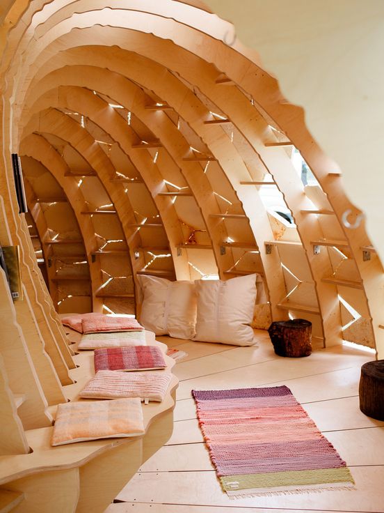 Photo: View into a cave-shaped, semi-circular wooden building with a bench, a carpet and cushions. Daylight streams in from the outside through gaps.