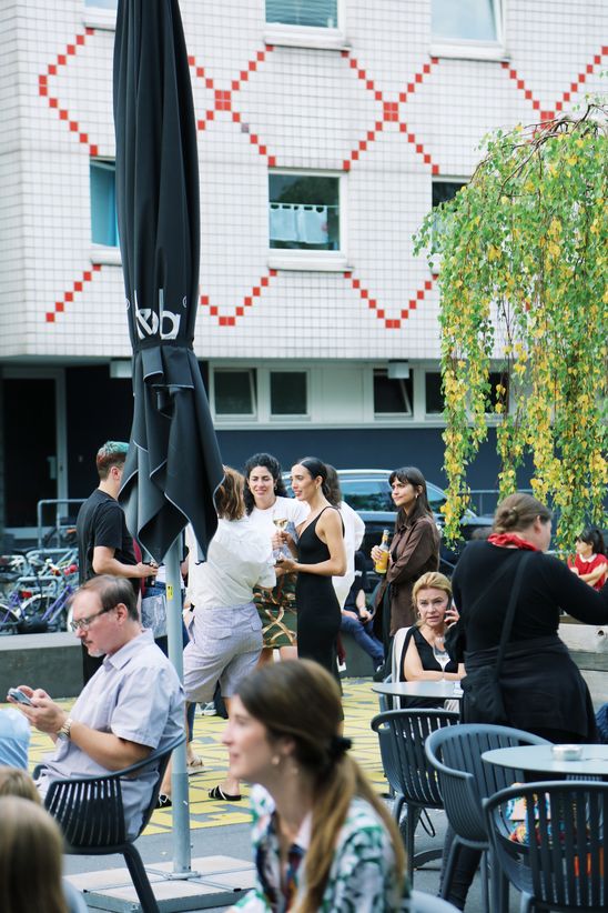 A couple of people are sitting around tables or standing, in the background a building with white and red tile facade.