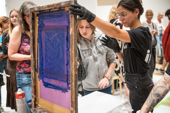 Photo: Several people stand around a screen printing station, one person holds up the screen printing frame and explains.