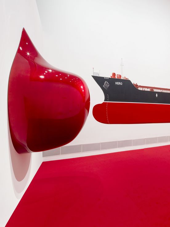 Photo: View of the exhibition room with its bright red carpet, a large reddish, shiny sculpture on the wall and a large painted oil tanker.