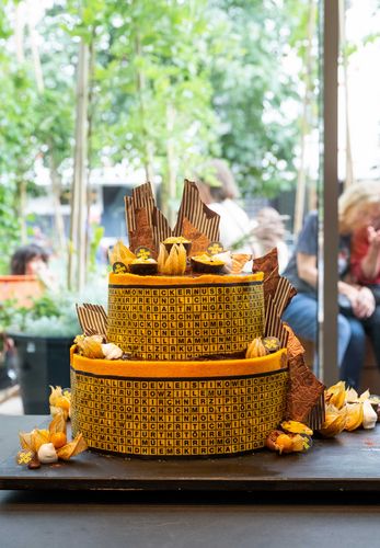 BG Geburtstagstorte von Sarah Klausen aus dem 180 Grad Cafè in Berlin Foto: Blick auf eine zweistöckige Geburtstagstorte, die mit braunen Buchstaben auf gelbem Untergrund, verschiedenen orangenen Früchten, Gold und Schokoladen verziert ist.
