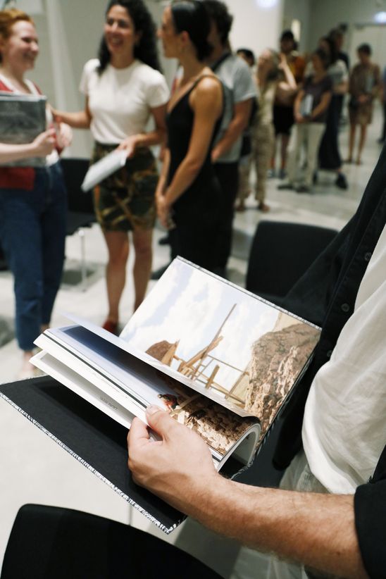 A person is flipping through the pages of an exhibition catalogue while a group of people is standing in the background chatting with each other.