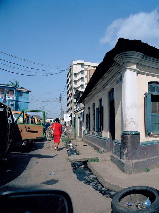 Square photograph: View of the front corner of a light yellow building with green shutters. To the right, a person in a red dress is walking along a narrow street.
