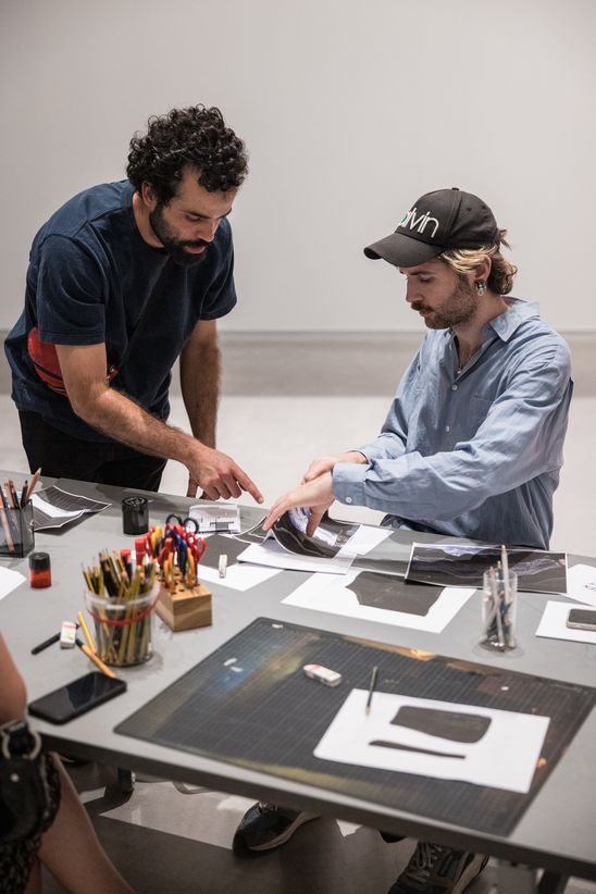 Photo: One person sits at a table and holds a sheet of paper in their hands, while another person stands slightly bent over next to them and explains something.