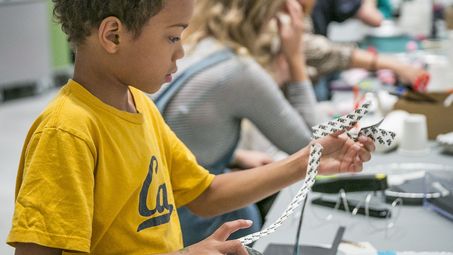Photo: A child is standing at a table with many craft materials. He is tearing strips off a tape roll.