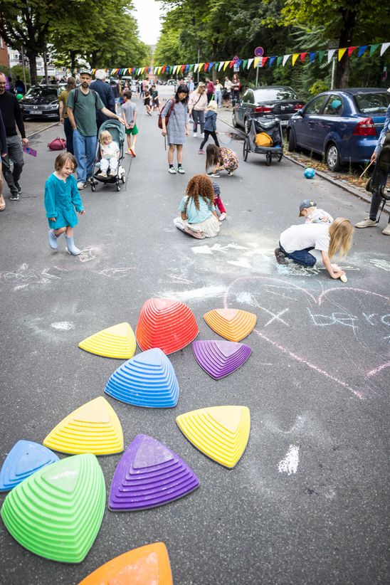 Photo: Many children play and paint on a cordoned-off street, over which colourful garlands have been hung.