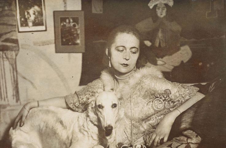 Selbstportrait der Künstlerin Marta-Astfalck-Vietz als Sepia-Fotografie. Sie sitzt in einem glänzenden Kleid mit Pelzbesatz auf einem Sofa. Auf ihrem Schoß sitzt ein Hund mit hellem Fell. Im Hintergrund sind Gemälde und dekorative Elemente zu sehen.