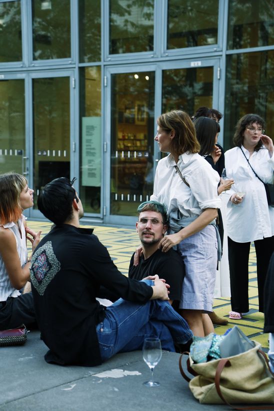 A group of people are sitting outside, a standing person has placed her hands on one of the seated people while looking to the side.