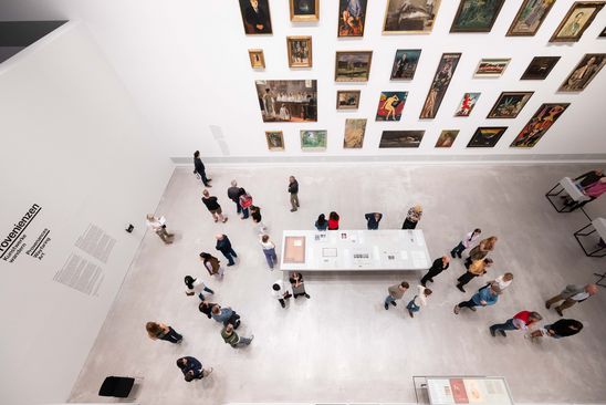 Photo: View from above of some people in the provenance exhibition, looking at works of art on the wall and archive material on a table in the room.