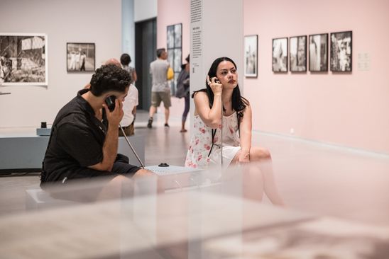 Photo: Two people sit at a listening station in the exhibition room and each hold a loudspeaker to their ear.