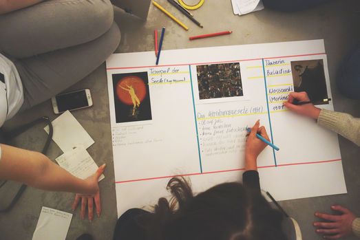 Foto: Verschiedene Hände notieren etwas zu drei verschiedenen Kunstwerken auf einem großen Papierbogen.