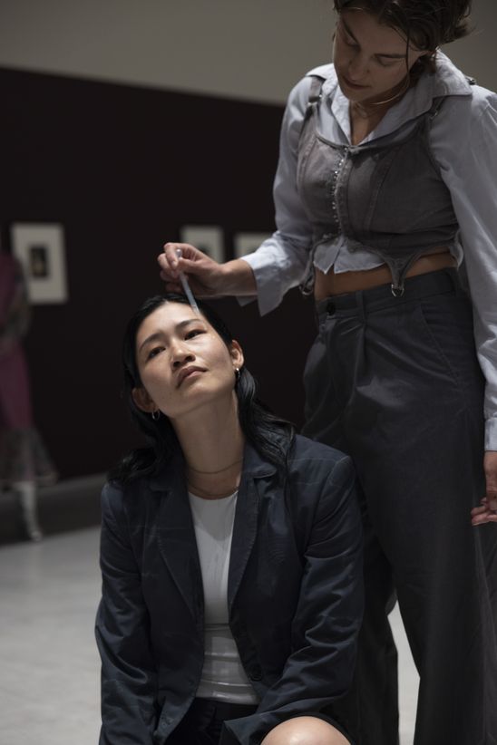 Foto: Deva Schubert steht hinter Chihiro Araki, die auf dem Boden sitzt, und tropft ihr mit einer Pipette Tränen auf die Wange.