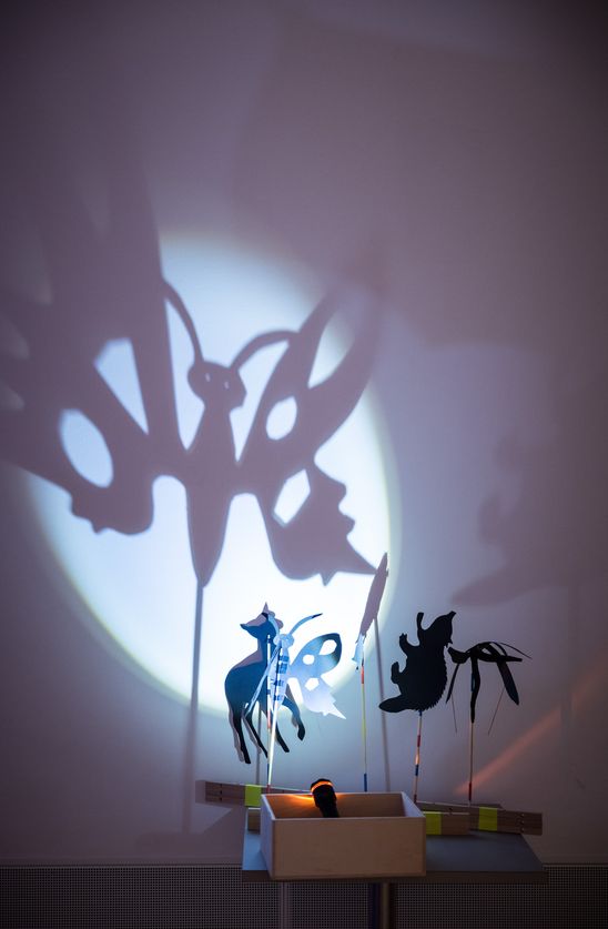 Photo: Two paper figures stand on a table and are illuminated from below with a torch. This creates a shadow play on the wall behind them.