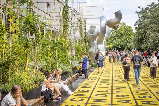 Photo: People sit along the forecourt of the Berlinische Galerie on a wooden platform between large planters and a silver sculpture or walk across the forecourt.