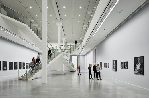 Foto: Menschen in einem zweistöckigen Ausstellungsraum mit Kunstwerken an den Wänden, in der Mitte zwei sich kreuzende Treppenaufgänge.