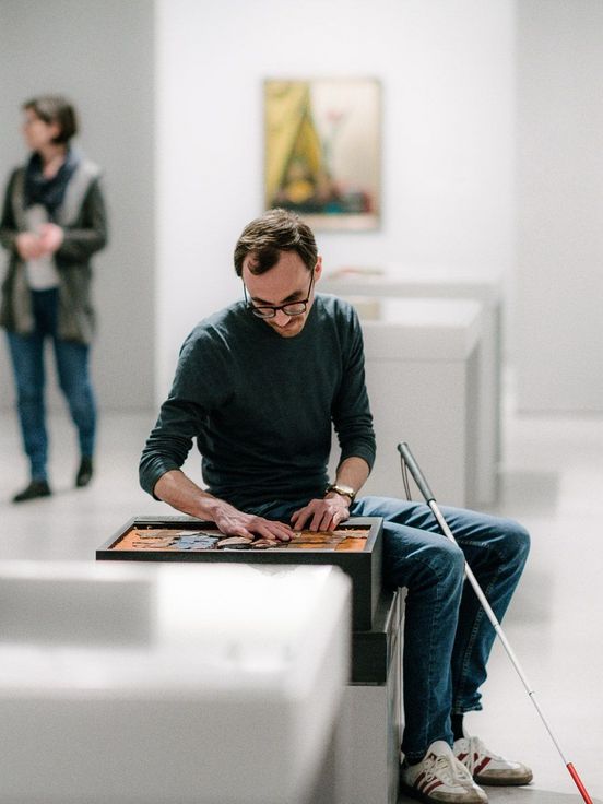 Foto: Besucher auf Sitzgelegenheit mit Blindenlangstock betastet ein Tastmodell im Ausstellungsraum.