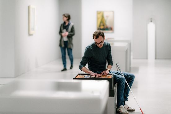 Foto: Besucher auf Sitzgelegenheit mit Blindenlangstock betastet ein Tastmodell im Ausstellungsraum.