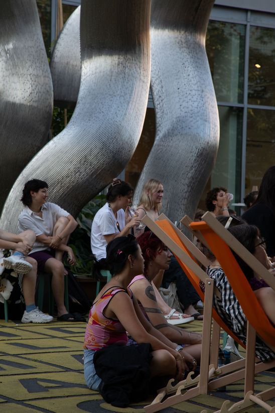 Foto: Seitlicher Blick auf einige Personen, die vor einer großen silbernen Skulptur auf dem Vorplatz der Berlinischen Galerie sitzen.