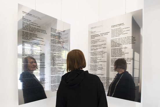 Foto: Käthe Kruse steht mit dem Rücken zur Kamera zwischen zwei großen, mit schwarzem Text bedruckten Spiegeln.