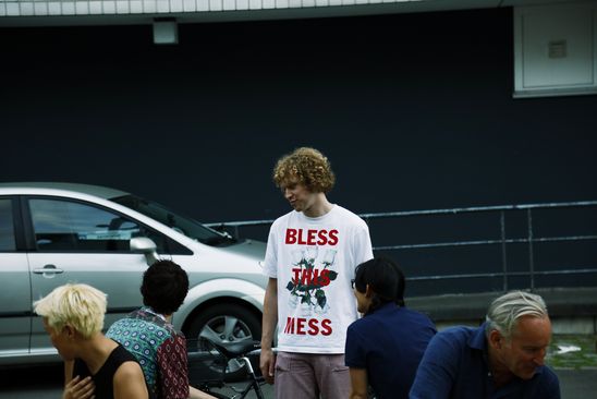A person is standing outside of the museum and chatting while other people are sitting. The person's white shirt reads in red font: "Bless This Mess". 