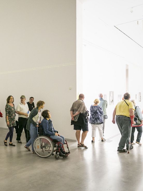 Foto: Viele Menschen unterschiedlichen Alters, mit und ohne Gehbehinderungen laufen durch einen Ausstellungsraum mit einer Laserinstallation.