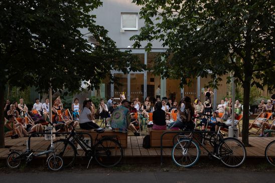 Foto: Viele Menschen sitzen auf bunten Stühlen, Hockern oder auf dem Boden auf dem Vorplatz der Berlinischen Galerie und lauschen einem Podium aus fünf Personen.