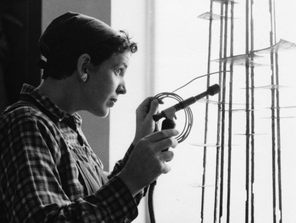 Schwarz-Weiß-Foto: Seitliche Aufnahme von Brigitte Meier-Denninghoff, die vor einem Fenster mit Lötkolben in der Hand an einer ihrer Skulpturen arbeitet.