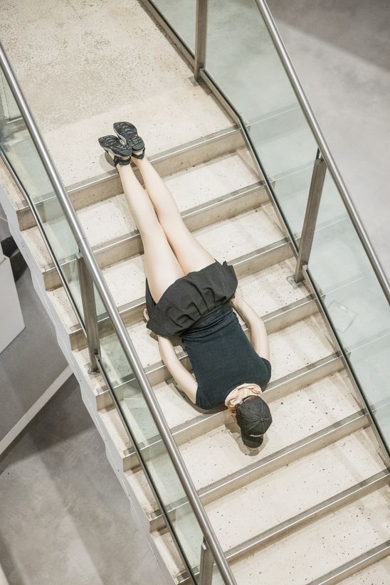 Photo: View from above of a person in a black dress with a cream-yellow top and tights, sliding down a flight of stairs on her stomach.