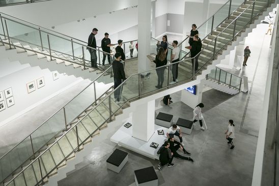 Foto: Viele Jugendliche stehen und sitzen verteilt in einem großen Raum mit zwei Treppen. Zuschauer*innen sehen ihnen bei der Performance zu.