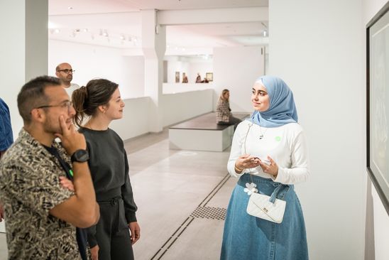 Photo: A schoolgirl stands in front of a framed work of art and talks to two visitors.