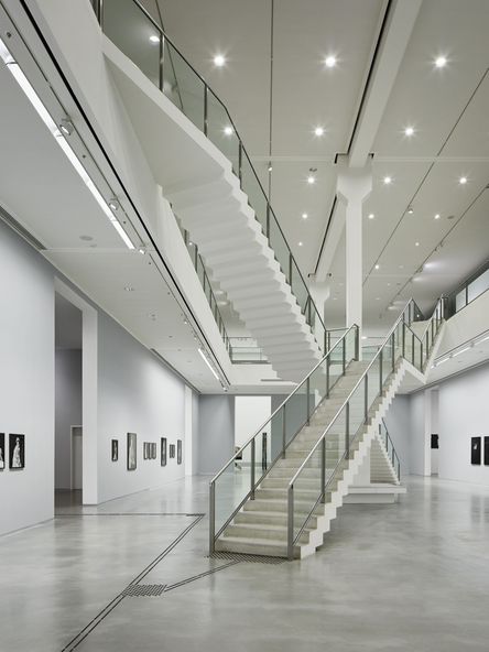 Foto: Zweistöckiger Ausstellungsraum mit Kunstwerken an den Wänden, in der Mitte zwei sich kreuzende Treppenaufgänge.