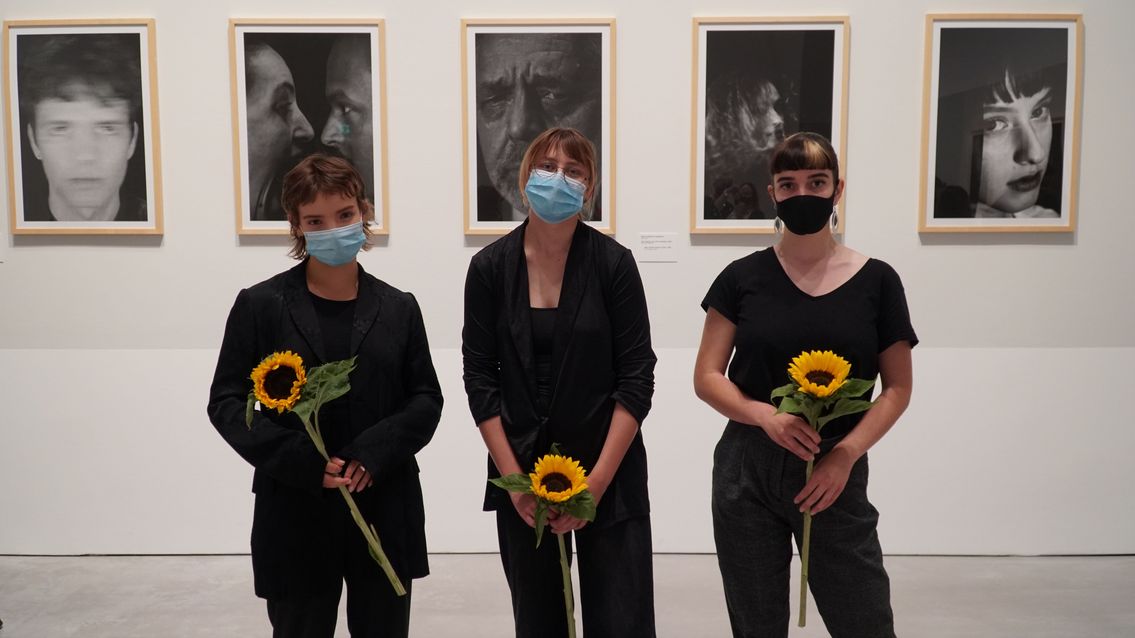 Foto: Drei junge Frauen stehen mit Blumen in der Hand vor Schwarz-Weiß-Fotografien.