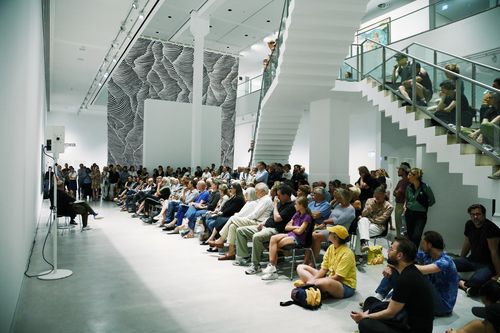 Impressions Book Launch and Artist Talk with Julius von Bismarck A large group of people is either sitting or standing in the exhibition space, in parts on the staircase crossing the room, while listening to a conversation.
